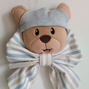Può includere: Una testa di orsacchiotto di peluche con un berretto di maglia azzurra e un papillon a righe. L'orso ha la pelliccia marrone, il naso rosa e gli occhi neri. Il papillon presenta strisce blu, beige e grigie.