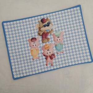 Può includere: Un tovaglietta a quadretti blu e bianchi con un lupo di cartone animato che indossa occhiali da sole e un cappello rosso, e quattro maiali di cartone animato che indossano cappelli. Il lupo è in piedi al centro della tovaglietta, e i maiali sono in piedi intorno a lui.