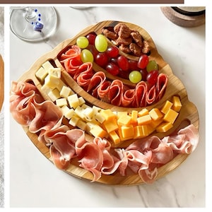 May include: A wooden serving board filled with an assortment of food items. The board contains sliced meats, cubed cheese, and grapes. Candied nuts are also present. The board is on a white marble surface.