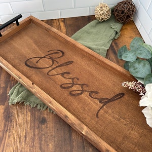 May include: A wooden serving tray with a dark brown stain and black handles. The tray has the word "Blessed" engraved on the surface.