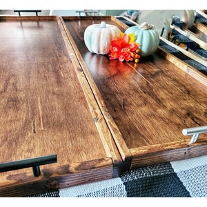 May include: Two wooden serving trays with black metal handles. The trays are stained a dark brown color and have a rustic look. The trays are sitting on a black and white checkered surface. There are two decorative pumpkins on the top tray, one orange and one blue.