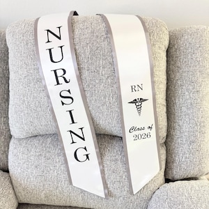 May include: White nursing sash with gray trim, featuring the word "NURSING" in black capital letters. The other side displays "RN", a caduceus symbol, and "Class of 2026". The sash is draped over a gray upholstered surface.