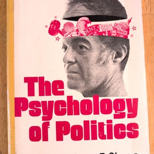 May include: Book cover for "The Psychology of Politics" by William F. Stone. The title is in large, bright pink letters. The cover features a black and white image of a man with a graphic of political symbols above his head.