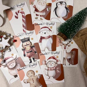 Könnte beinhalten: Neun weihnachtliche Bügelbilder mit verschiedenen Figuren, darunter Weihnachtsmann, Pinguin, Eisbär, Nussknacker, Rentier, Lebkuchenmann, Maus, Yak und Zuckerstange. Die Bügelbilder sind auf weißen Karten mit braunen und rosafarbenen Akzenten und dem Text "Schneck BERT Bügelbild".