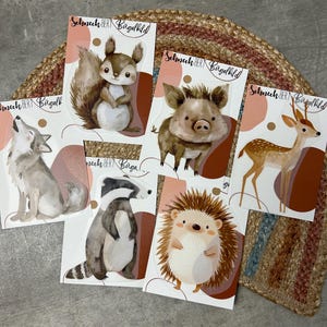 Könnte beinhalten: Sechs Bügelbilder mit Aquarell-Illustrationen von Waldtieren. Die Tiere sind ein Eichhörnchen, ein Eber, ein Reh, ein Wolf, ein Dachs und ein Igel. Die Bügelbilder sind auf weißem Hintergrund mit einem rosa und braunen Design.
