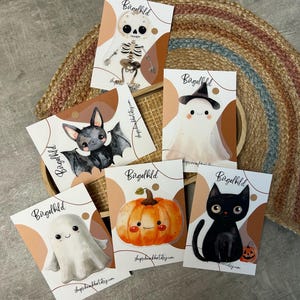 Könnte beinhalten: Eine Sammlung von Halloween-Karten mit Illustrationen eines Skeletts, einer Fledermaus, eines Geistes, eines Kürbisses und einer schwarzen Katze. Jede Karte trägt das Wort "Bügelbild" und eine Shop-Website-Adresse.