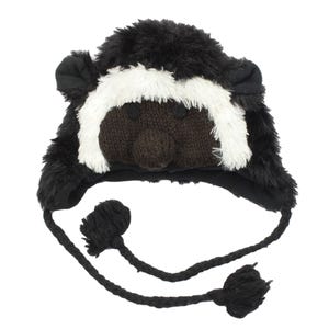 May include: Black and white furry hat with a brown knitted face and black pom poms on the ends of the ties.