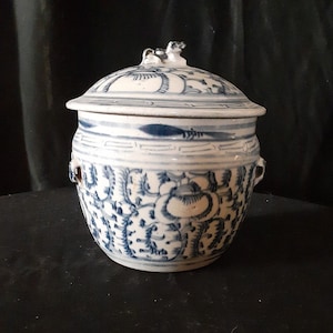 Puede incluir: Un tarro de cerámica blanca con un diseño floral azul y blanco. El tarro tiene una tapa con una pequeña figura de animal en la parte superior.