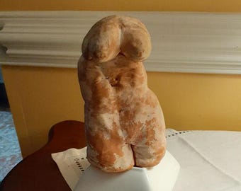 May include: A terracotta sculpture of a nude female figure with a rounded head and arms crossed over the chest. The sculpture is standing on a white square base.