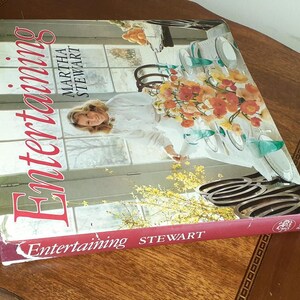 May include: A red hardcover book titled "Entertaining" by Martha Stewart. The cover features a woman in a white shirt and a floral arrangement on a table with plates and glasses.