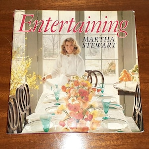 May include: A book titled "Entertaining" by Martha Stewart, featuring a photo of a woman in a white shirt standing in a dining room with a table set for a meal. The table is decorated with flowers and there are chairs around the table.