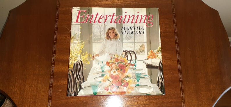 May include: A book titled "Entertaining" by Martha Stewart, featuring a photo of a woman in a white shirt standing in a dining room with a table set for a meal. The table is decorated with flowers and there are chairs around the table.