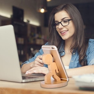 Może przedstawiać: Drewniany stojak na telefon w kształcie osoby trzymającej smartfon. Stojak wykonany jest z jasnego drewna i ma zaokrągloną podstawę. Smartfon jest w kolorze złotym.