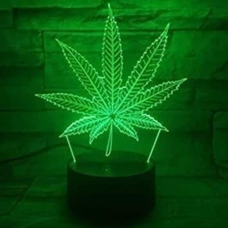 May include: Green LED night light shaped like a marijuana leaf. The light is on and illuminating the leaf.