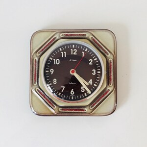 Könnte beinhalten: Eine quadratische Wanduhr im Vintage-Stil mit schwarzem Zifferblatt und weißen Ziffern. Die Uhr hat einen roten Sekundenzeiger und cremefarbene Stunden- und Minutenzeiger. Das Zifferblatt ist von einem cremefarbenen Rahmen mit roten Akzenten umgeben. Das Wort "Quartz" ist aufgedruckt.