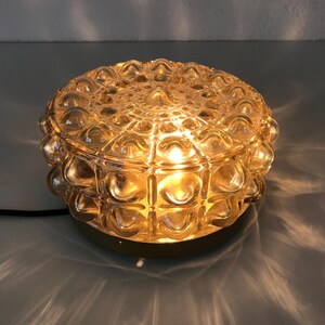 Könnte beinhalten: Eine runde Vintage-Tischlampe mit einem strukturierten, blasenartigen Glasschirm. Die Lampe strahlt ein warmes, goldenes Licht aus und wirft interessante Schatten. Der Sockel hat eine hellgoldene Farbe. Die Lampe ist mit einem schwarzen Netzkabel verbunden.