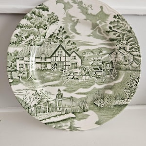 May include: A round, white ceramic plate with a green transferware design. The scene depicts a rural landscape with buildings, trees, a horse-drawn carriage, and figures. The plate has a scalloped edge.