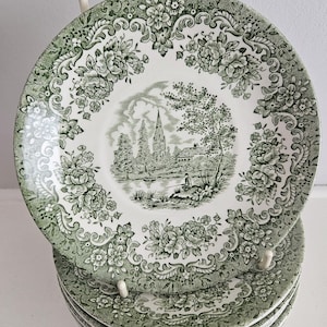 May include: A stack of round ceramic plates featuring a green and white transferware pattern. The center depicts a landscape with a church spire, trees, and a person fishing by a lake. The wide border displays intricate floral and scroll motifs.