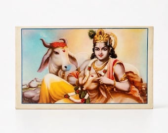May include: A rectangular print featuring a figure with dark hair, a golden crown with a peacock feather, and gold jewellery, wearing orange and red garments. The figure is seated beside a light-coloured cow, with a soft, warm-toned background.