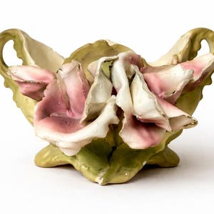 Può includere: Una ciotola in ceramica con due manici laterali e un elemento decorativo centrale composto da petali di rosa scolpiti e sovrapposti in tonalità bianco e rosa tenue. La base e i manici presentano una finitura verde oliva opaca.