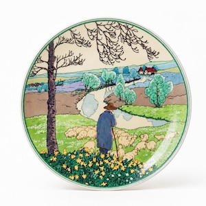 Könnte beinhalten: Ein runder Keramikteller mit einer farbenfrohen Illustration eines Hirten im blauen Mantel, der mit einer Schafherde auf einem Feld mit gelben Blumen steht. Die Szene zeigt einen gewundenen Fluss, grüne Bäume und ein entferntes Haus.