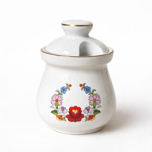 Puede incluir: Un azucarero de cerámica blanca con tapa que incluye una pequeña muesca para una cuchara. El cuerpo del recipiente está decorado con una corona floral colorida en tonos rojos, rosas, azules, amarillos y verdes. La tapa y el borde tienen un filo dorado.