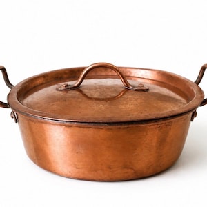 Puede incluir: Una olla redonda de cobre con tapa a juego y dos asas laterales. La olla presenta un acabado metálico cálido con signos visibles de uso y un cuerpo curvo y liso. La tapa tiene un asa central arqueada y las asas laterales están fijadas.