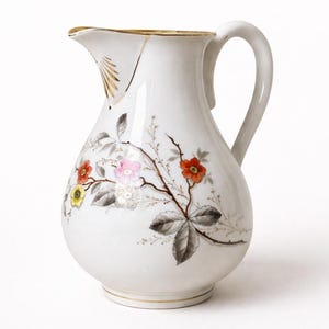 Peut inclure: Un pichet en porcelaine blanche orné d'un motif floral peint à la main avec des fleurs rouges, roses et jaunes sur des branches brunes aux feuilles grises. Le pichet possède une anse courbée, un bec verseur et un liseré doré sur le bord.