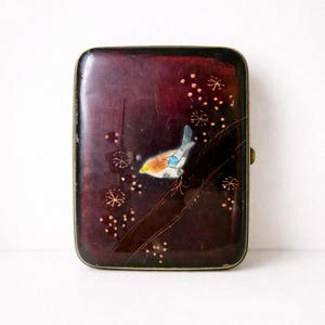 Può includere: Un astuccio rettangolare vintage con sfondo bordeaux scuro e un disegno dipinto di uccelli e fiori. L'uccello ha accenti gialli, arancioni e blu. L'astuccio ha una cornice e una chiusura in metallo color oro.