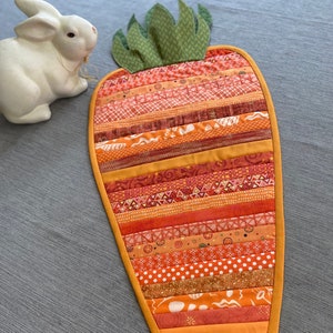 May include: A handmade carrot-shaped quilted table runner. The runner features various orange and red fabrics in horizontal strips, with a green fabric top. The edges are trimmed with a yellow-orange binding. A white ceramic bunny is in the background.