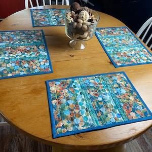 Puede incluir: Un juego de tres manteles individuales rectangulares acolchados con un patrón vibrante de círculos multicolores en tonos azul, naranja y verde. Cada mantel está acabado con un borde de tela azul sólido y costuras verticales en la superficie.