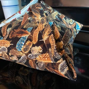 May include: A square, quilted fabric bowl cozy with a black background and a pattern of brown, tan, and blue feathers. The cozy is folded and appears to be on a black surface.