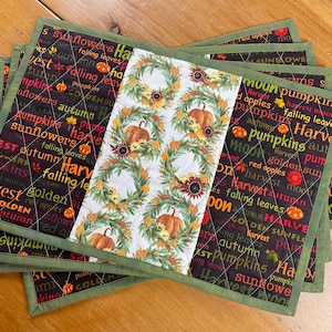 May include: A set of rectangular quilted placemats featuring a central white panel with a wreath, pumpkin, and sunflower print, flanked by black side panels printed with autumn-themed words like pumpkins, sunflowers, and harvest in orange and yellow.