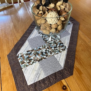May include: A glass bowl filled with decorative dried botanicals sits on a quilted table runner. The runner features a patchwork design with brown, gray, and blue squares, framed by a dark brown wood-grain border. The table is made of wood.