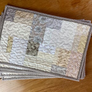 Puede incluir: Juego de seis manteles individuales acolchados en beige y blanco con un diseño de patchwork. Los manteles individuales tienen un borde decorativo.