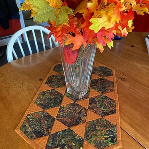 Puede incluir: Un camino de mesa marrón con un patrón geométrico de hexágonos. El camino de mesa está hecho de tela con un estampado paisley en tonos de verde, marrón y naranja. Un jarrón con hojas de otoño en tonos de naranja, amarillo y rojo se encuentra sobre el camino de mesa.