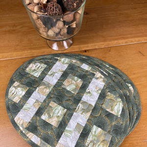 May include: A set of oval quilted placemats featuring a patchwork design with alternating squares of green and white fabric. The green fabric has a gold dotted pattern, while the white fabric displays a repeating print of light green pumpkins.