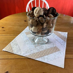 May include: A clear glass pedestal bowl filled with decorative items sits on a quilted placemat. The placemat features a patchwork design with light beige, white, and floral patterned fabric. The bowl contains various nuts and decorative wooden pieces.