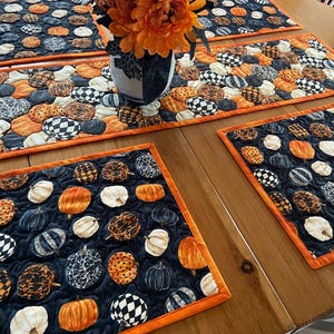 May include: A set of quilted fabric placemats and a table runner featuring a repeating pattern of pumpkins in orange, black, white, and grey with various textures and prints, finished with a bright orange fabric border trim on all edges.