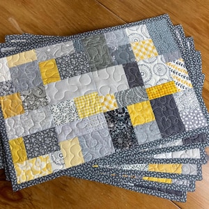 May include: A rectangular quilted placemat featuring a patchwork design of various fabric squares in shades of gray, white, and yellow. The fabric patterns include gingham, floral, and geometric prints, all joined with visible stippled quilting stitches.