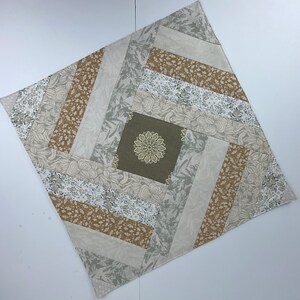 May include: A square quilted placemat with a central olive green square featuring a floral design. The placemat has a log cabin design with alternating strips of beige, gray, and brown floral patterns.