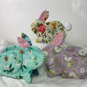 Peut inclure: Trois pochettes en forme de lapin en tissu avec des motifs floraux. L'une est vert menthe, l'autre lavande et l'autre crème. Chacune a des oreilles intérieures roses et une queue en pompon blanc. Les pochettes sont matelassées.