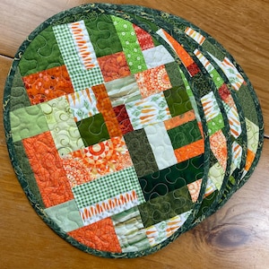 May include: Set of four round quilted placemats with a patchwork design. The placemats feature a variety of green, orange, and white fabrics with carrot and floral patterns. Each placemat has a dark green border.