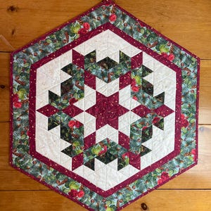 May include: A quilted, hexagonal table runner with a festive design. The center features a starburst pattern in white and burgundy, surrounded by a border of floral and pine needle fabric. The outer edge is a deep burgundy.
