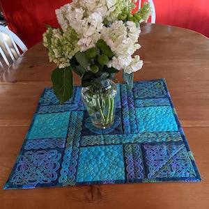 May include: A square, quilted table runner with a Celtic knot design in shades of blue and teal. The runner is decorated with a variety of patterns and textures. A glass vase with white and green flowers sits on top of the runner.