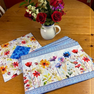 Puede incluir: Un ramo de rosas rojas y flores blancas en una jarra blanca con borde azul sobre una mesa de madera. Dos manteles individuales florales con borde azul y flores coloridas están sobre la mesa.