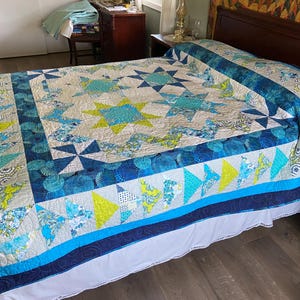 Puede incluir: Un edredón hecho a mano con un diseño de estrella y molinillo en tonos de azul, verde azulado y amarillo. El edredón está sobre una cama con una falda de cama blanca. El borde del edredón presenta una mezcla de patrones y colores.