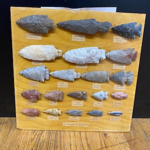 May include: A wooden display board featuring a collection of various arrowheads, each labeled with a state name. The arrowheads are in different colors and shapes, mounted on the wooden surface. The text "Indian Spear and Arrowheads" is at the bottom.