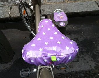 May include: A purple bicycle seat with white stars, a brown bicycle frame, and a black handlebar. The bicycle is parked on a cobblestone street. The seat has a white star pattern and a green tag.