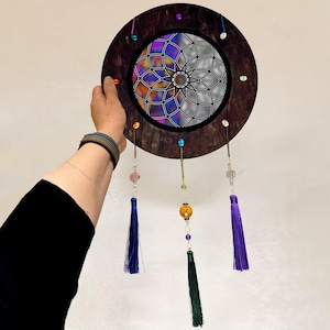 May include: A wooden circular dreamcatcher with a colorful stained glass mandala design. It has three purple, green, and blue tassels hanging from it.